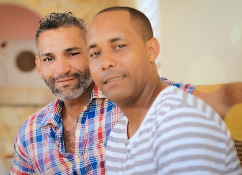 Portrait of homosexual couple, happy gay people smiling at camera, sitting on sofa at home. Same sex marriage between hispanic men.